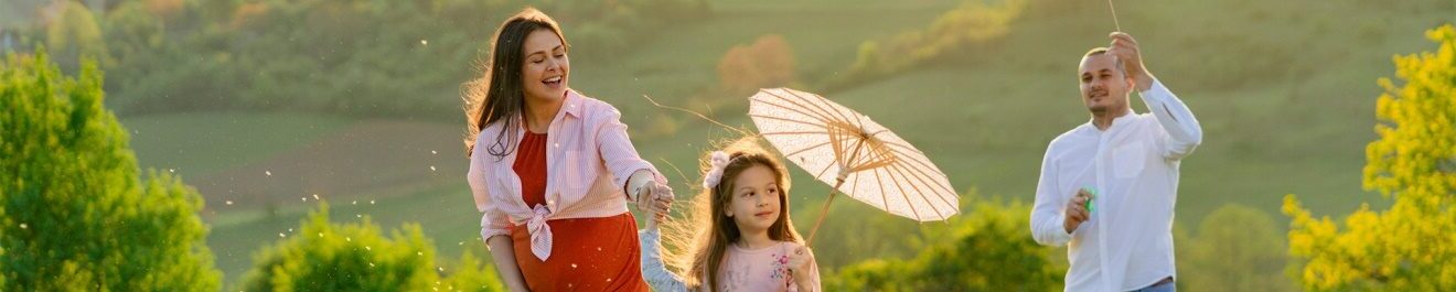 Une famille marchant dans un paysage avec la femme enceinte, la fille tenant un parapluie et le père un cerf volant.